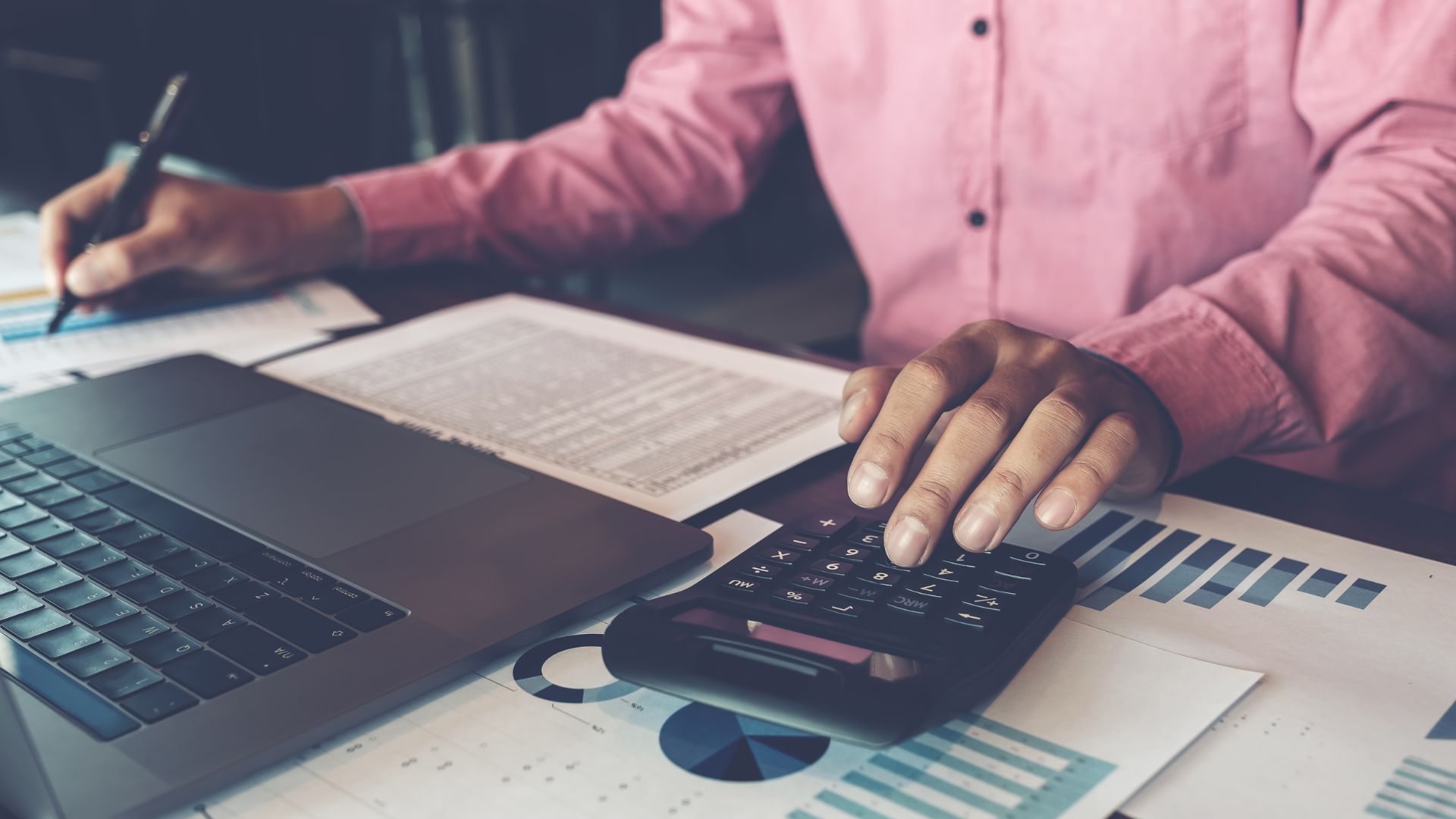 Professional person with pink shirt sitting at desk in front of laptop, working with calculator and various papers, a standard sight in bookkeeping for attorneys