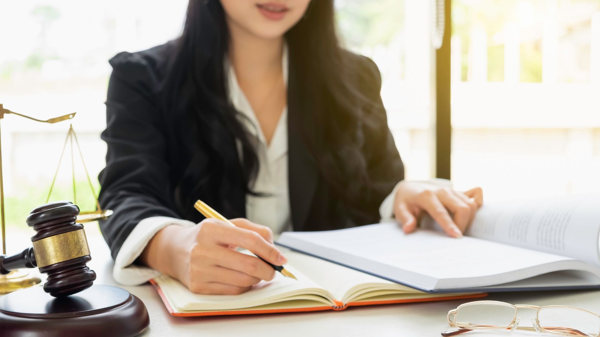 a legal expert in liquidation looking over ledgers at a desk, pen in hand, there is a gavel and scales on one side of the desk and a pair of glasses on the other,