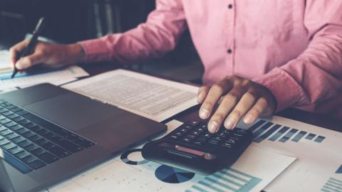 Professional person with pink shirt sitting at desk in front of laptop, working with calculator and various papers, a standard sight in bookkeeping for attorneys